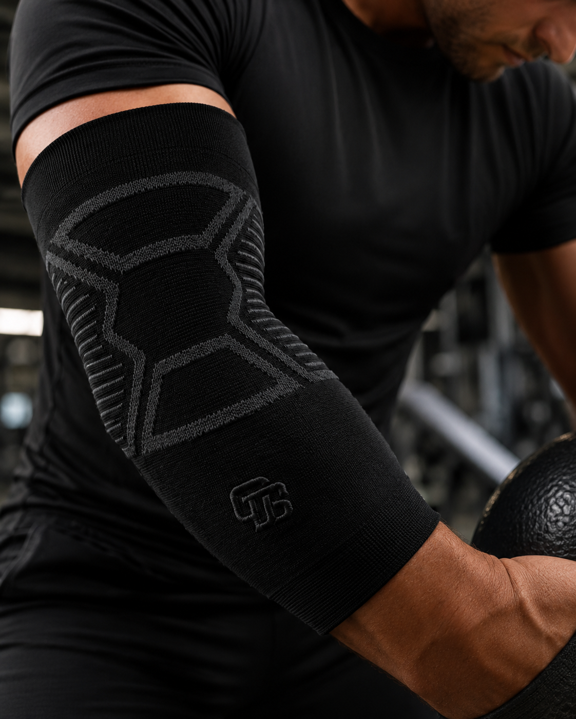 Person wearing a black elbow support with a logo, holding a medicine ball in a gym setting.