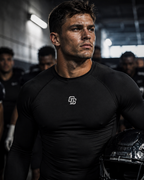 Man wearing a black athletic shirt with a logo, holding a helmet in a dark indoor setting.