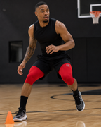 Person wearing black sleeveless shirt and shorts with red knee sleeves on a basketball court.