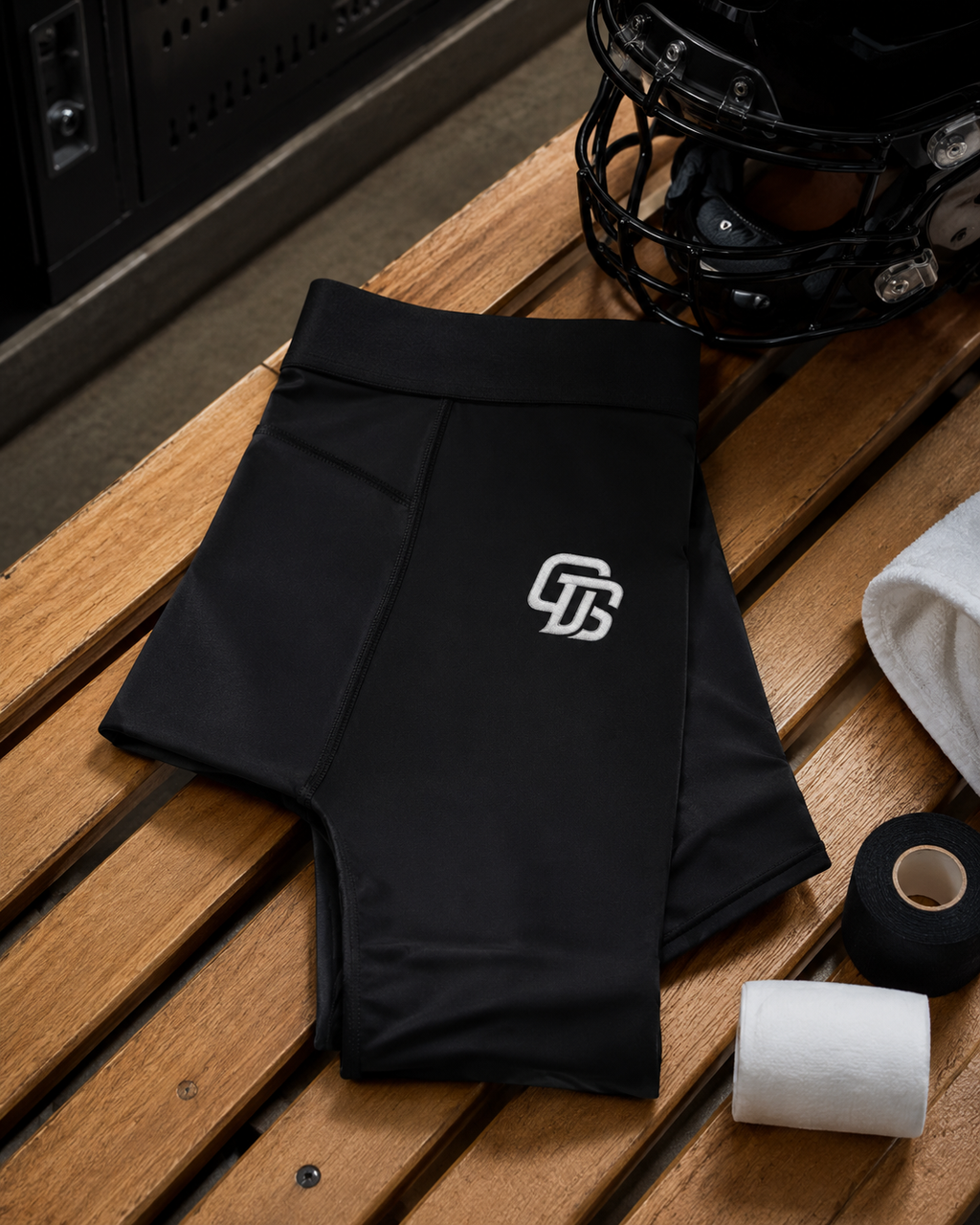 Black athletic shorts with a logo on a wooden surface, next to a helmet and tape.